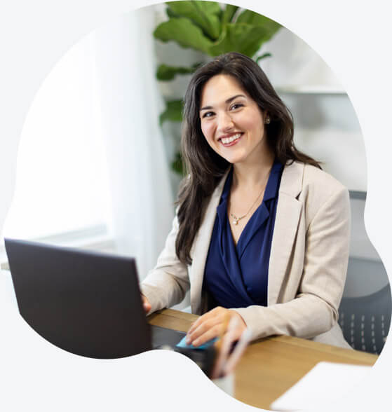 Woman at her office wearing a blazer and working on a laptop while smiling at camera