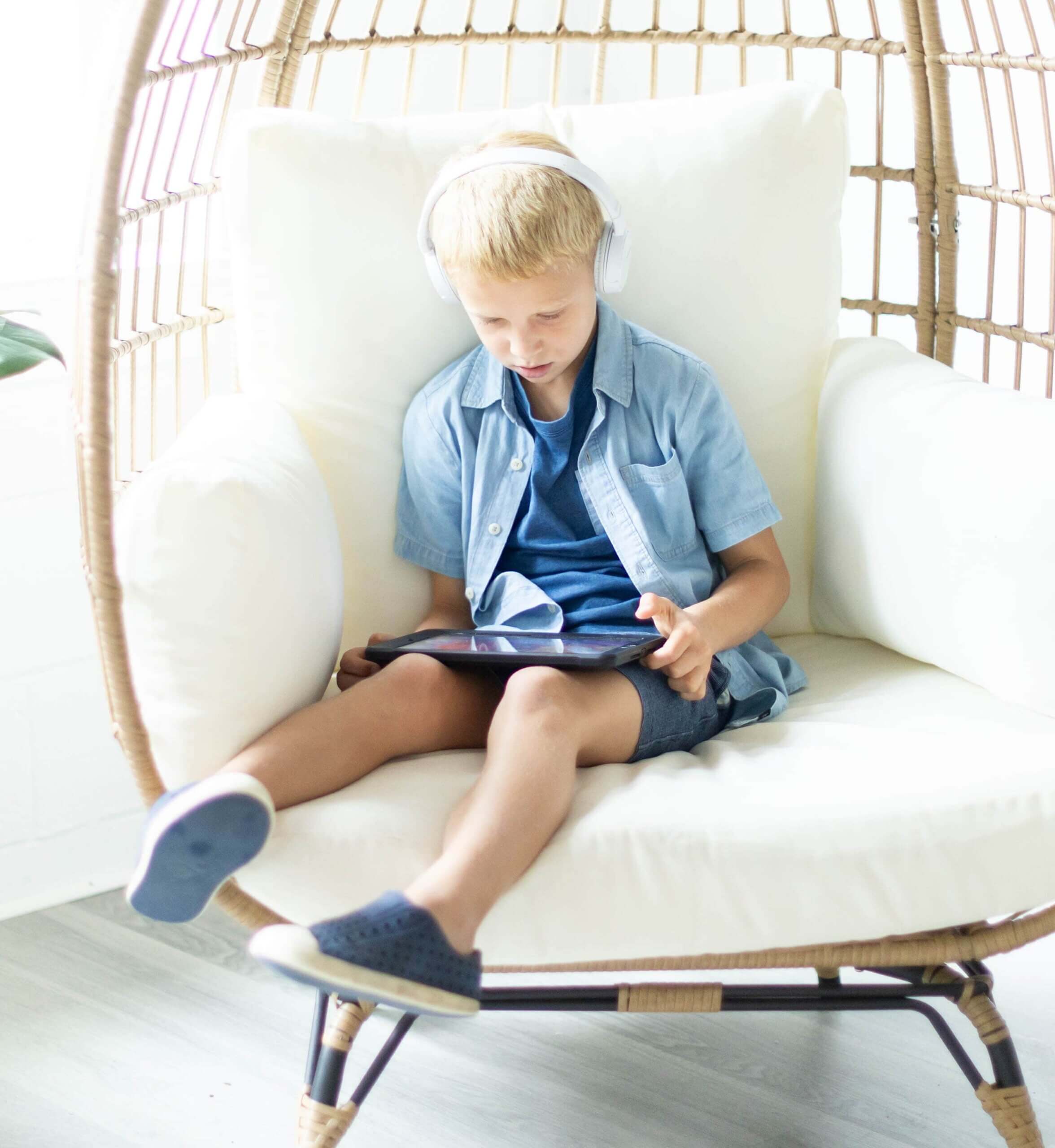 Little boy sitting on chair with headphones on and a tablet in their hands